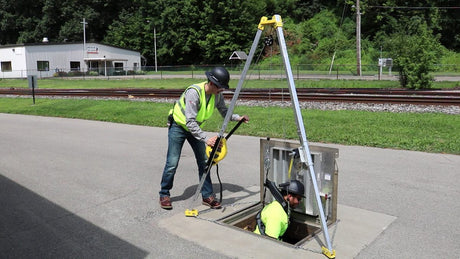 One worker lowering another into a manhole using a tripod, harness and other fall safety equipment.