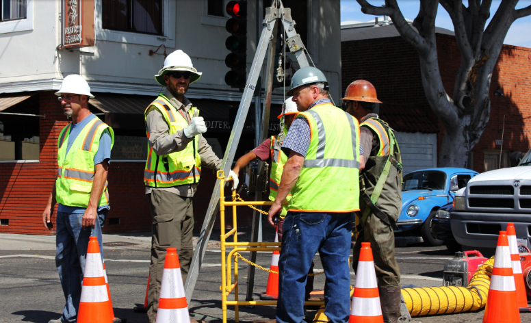 Utility workers standing on a street with tripod and ventilation over a man hole. Workrs wearing hi-vis vests, hard harts and safety cones are placed around them.