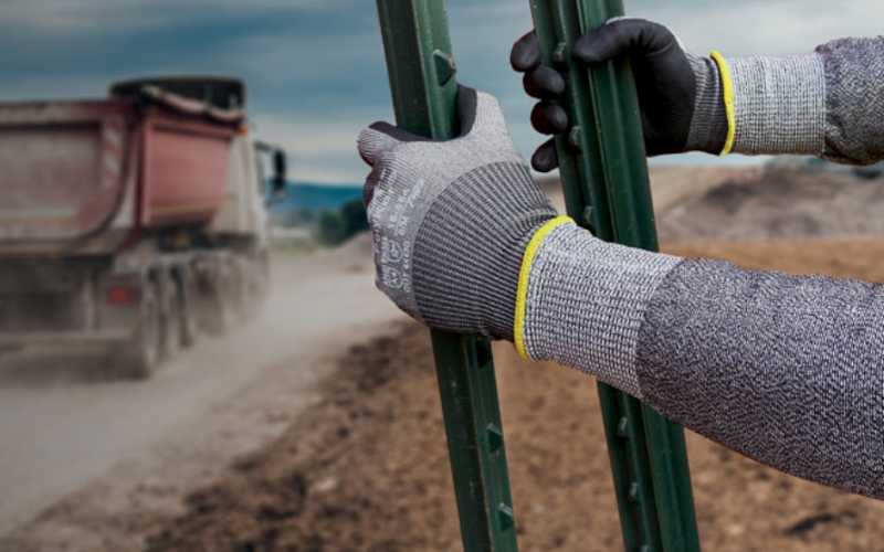 Worker wearing protective gloves holds a metal beam on a construction site. A dump truck drives by, raising dust, under a cloudy sky.