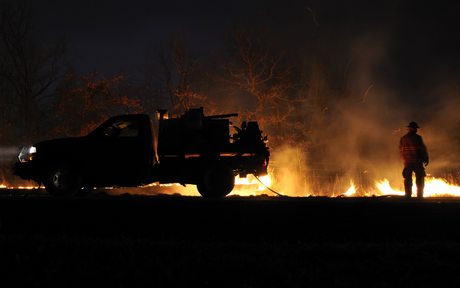 A silhouetted firefighter stands beside a truck at night, battling a controlled fire with glowing embers and smoke rising in the dark, dramatic scene.
