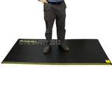 A person stands on a large, black Working Concepts ErgoKneel Extreme Standing Mat with yellow-striped edges. Only the lower body is visible. The mat features text indicating that it's a product from PK Safety 2012.