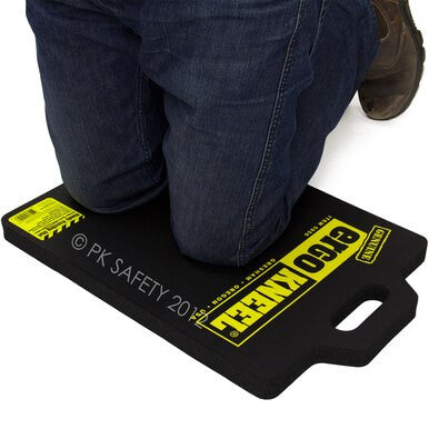 A person in jeans and work boots kneels on a Working Concepts Large ErgoKneel Kneeling Mat 5050, measuring 14 x 21 inches. The ergonomic mat is made from black closed-cell foam and features yellow text that reads "Pro Knee," along with essential safety information. Thanks to its handle, it is convenient to carry.