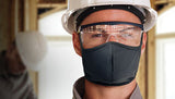 A construction worker wearing a hard hat, safety goggles, and a black face mask stands indoors at a building site, equipped with Howard Leight Laser-Lite Disposable Corded Earplugs for hearing protection. Another worker is blurred in the background, also wearing a hard hat and prioritizing noise reduction using Howard Leight earplugs.