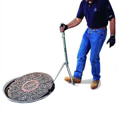 A person in jeans and a dark shirt uses the Allegro Manhole Lid Lifter Steel Handle 9401-20, equipped with rubber grips, to lift the patterned cover. Wearing gloves and boots, they stand confidently on a white background, showcasing the tool's steel handle that enhances both strength and precision.