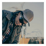 A person in a khaki cap, plaid shirt, jacket, and MCR Safety Law OTG Safety Glasses UV-AF Anti-Fog OG1 looks down outdoors on a sunny day, with a blurred fence and branches in the background.