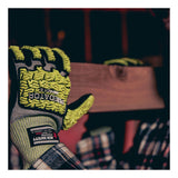 A person wears MCR Safety Predator Hi-Vis Mechanics Gloves w/HyperMax PD6952 (12 pairs) in green and black, holding a wooden plank as the cut-resistant gloves stand out in the dimly lit workshop.