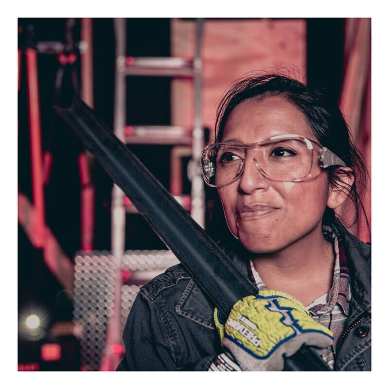 A woman wearing MCR Safety Law OTG Safety Glasses UV-AF Anti-Fog OG1 and work gloves holds a black metal bar, standing before a ladder and construction materials with a determined expression.