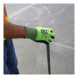 A person wearing a high-visibility vest and bright green MCR Safety CutPro HyperMax MicroFoam Nitrile Gloves (92785NF) holds a gray metal rod against a concrete floor. The gloves display "CUT PRO" branding and safety certification icons.