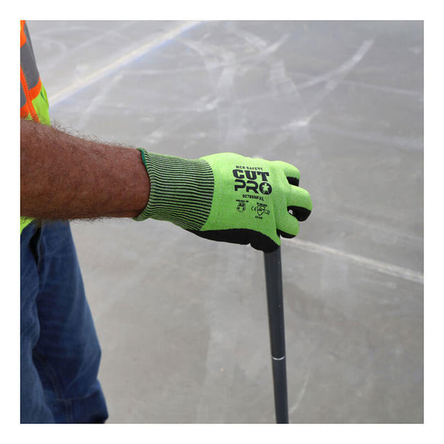 A person wearing a high-visibility vest and bright green MCR Safety CutPro HyperMax MicroFoam Nitrile Gloves (92785NF) holds a gray metal rod against a concrete floor. The gloves display "CUT PRO" branding and safety certification icons.