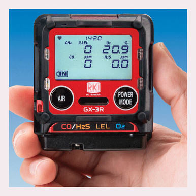 A person's hand holds the RKI Instruments GX-3R 4 Gas Confined Space Monitor 72-RA-C, a compact device equipped with micro sensors. The digital display shows levels for CH4, O2, CO, and H2S. Designed for confined spaces, it features buttons labeled "Air" and "Power Mode.