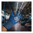 A gloved hand wearing a blue PIP G-Tek Paradox PolyKor A6 Foam Nitrile Glove 16-361 holds a small metal part in a factory setting, with machinery and equipment visible in the background.