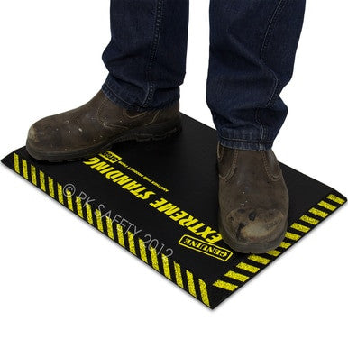 A person wearing brown work boots stands on a black Working Concepts ErgoKneel Extreme Standing Mat 14 x 21 in., featuring yellow caution stripes and the text "EXTREME STANDING" in bold, while utilizing an anti-fatigue mat for enhanced comfort.