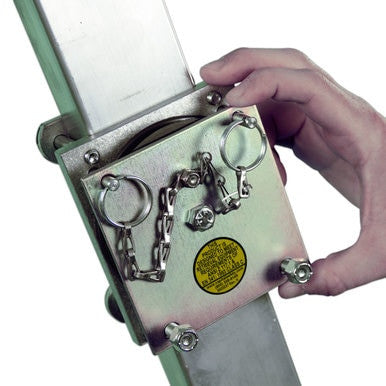 A person's hand adjusts a metallic safety lock mechanism on an extension ladder. This 3M DBI-SALA Fall Protection device utilizes a chain and pin system for securing the ladder sections. A yellow safety label with instructions is visible alongside the DBI-SALA Tripod Leg-Mount Pulley, model 8003238.