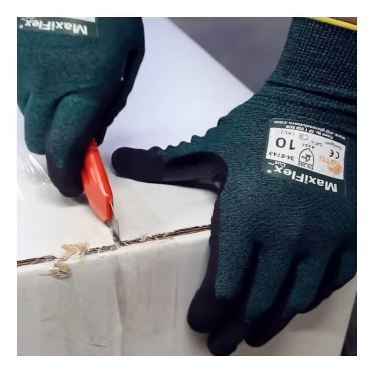A person wearing PIP ATG MaxiFlex Cut Resistant Glove 34-8743 uses a red utility knife to cut open a sealed cardboard box along the tape.