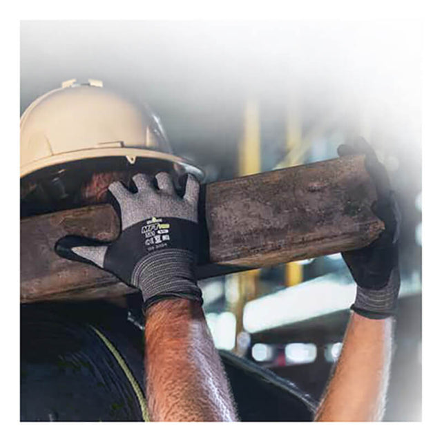 A construction worker wearing SHOWA 381 General Purpose Gloves by Showa Best and a hard hat carries a wooden beam on their shoulder, with a blurred industrial background.