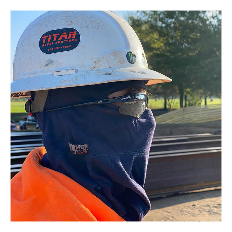 A construction worker in a white hard hat, dark safety glasses, orange jacket, and an MCR Safety FR Westex True Comfort Balaclava Navy BLCVTCN stands outdoors near stacked metal beams, with trees in the background.