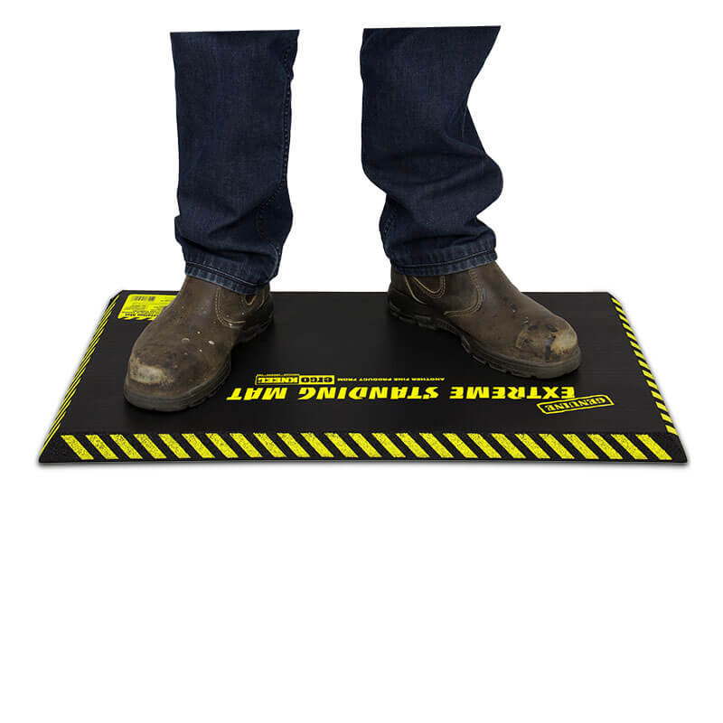 A person wearing muddy boots stands on the Working Concepts ErgoKneel Extreme Standing Mat 5020, measuring 16 x 28 inches—perfect for workers who need to stand for extended periods. This anti-fatigue mat offers a textured surface along with caution stripes around the edges, ensuring comfort and safety in challenging settings.