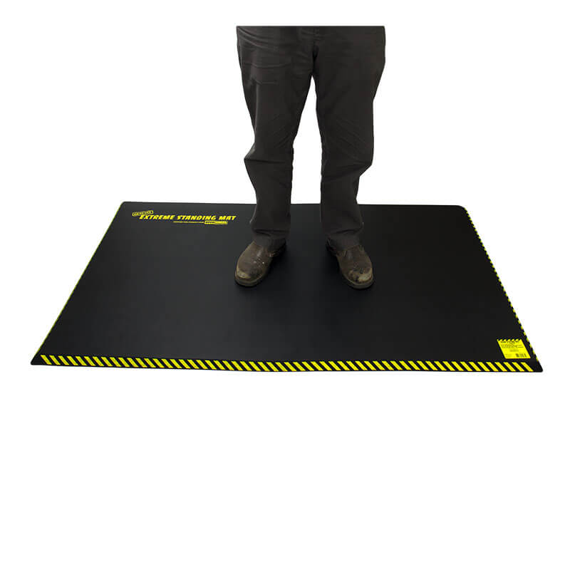 An individual stands on a Working Concepts ErgoKneel Extreme Standing Mat 5035, measuring 36 x 60 inches, featuring a black surface with yellow striped borders. Designed for support and comfort in extreme conditions, the mat is likely crafted from durable nitrile foam. The individual's face is not visible.