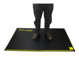 An individual stands on a Working Concepts ErgoKneel Extreme Standing Mat 5035, measuring 36 x 60 inches, featuring a black surface with yellow striped borders. Designed for support and comfort in extreme conditions, the mat is likely crafted from durable nitrile foam. The individual's face is not visible.