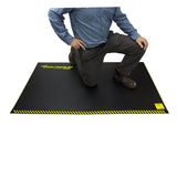 A person in a blue shirt and gray pants kneels on a Working Concepts ErgoKneel Extreme Standing Mat 5035, featuring yellow caution stripes and ensuring both safety and comfort during prolonged tasks.