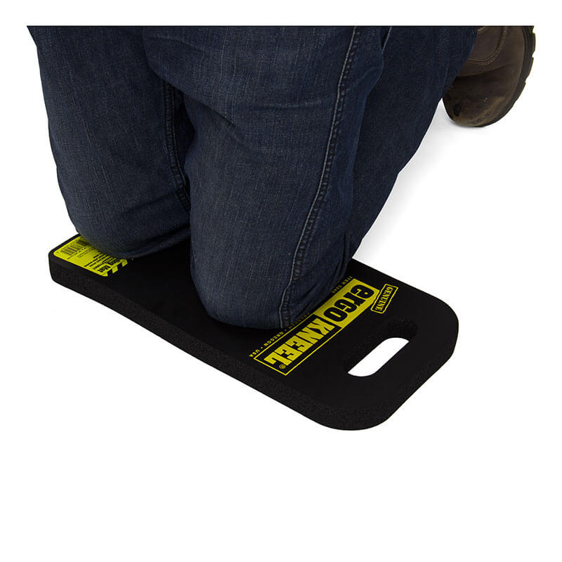 A person is kneeling on the ergonomic foam rubber mat labeled "Working Concepts ErgoKneel Kneeling Mat 5040 8 x 16 in." by Working Concepts. The black mat, with a handle cutout on one end, provides support for someone wearing dark blue jeans and brown work boots.