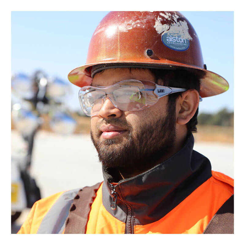 A construction worker wearing a rust-colored hard hat, MCR Safety Law OTG Safety Glasses UV-AF Anti-Fog OG1, and an orange high-visibility jacket stands outdoors on a sunny day.