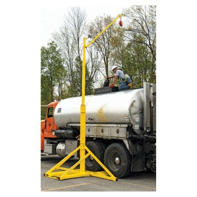 A worker is atop a tanker truck, clad in safety gear and adjusting equipment. Beside the truck, a portable yellow Miller SkyORB Rotational Boom Anchor SORBSYS from Miller Fall Protection provides secure overhead anchorage. In the background, trees and an orange truck add to the scene's bustling industrial atmosphere.