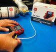 Hands are working with a red multi-gas personal monitor from RKI Instruments, model GX-2009, which is connected to a gas canister on a blue workbench. In the background, there is a white box featuring the portable gas monitor's image and RKI Instruments branding, suggesting its precision for gas detector calibration in line with the RKI Instruments Gas Monitor Calibration Service.