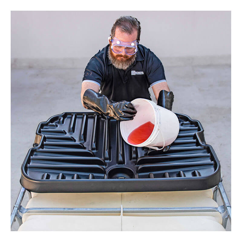 Donning safety goggles and gloves, a person uses the UltraTech IBC Funnel 0648 by UltraTech International to carefully transfer red liquid from a white bucket into a large black container. The container is securely positioned on a metal frame, highlighting the industrial setting's commitment to precision and chemical resistance.