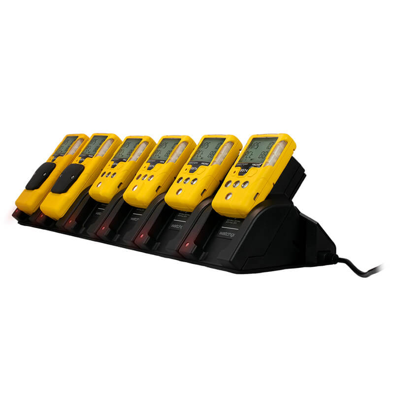 Six yellow WatchGas handheld gas detectors are neatly aligned in a row on the black WatchGas SST 6 Way Bench Top Charger (SST-6WSCC-KIT). Each device features a digital display and buttons, while the charger—designed as a centralized solution for charging up to six units simultaneously—is connected to a power source.