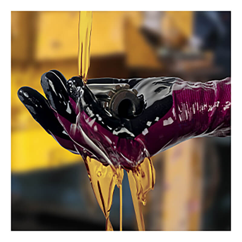 A gloved hand, protected by PIP ATG MaxiDry Elite Fully Coated Gloves 64-226, holds a small metal part as yellow oil pours over it, with blurred machinery in the background.
