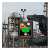 A gloved hand holds an RKI Instruments HS-04 Series H2S Single Gas Monitor 73-0063 with a digital display reading “0.0,” set against an industrial facility featuring metal structures and tanks in the background.