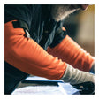 A person with a gray beard wears MCR Safety 13g Hi-Vis HyperMax Cut Pro Sleeves (92_ _OVT) and gray gloves while working with tools, focusing on a task at a workstation.