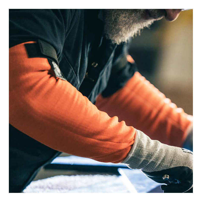 A person with a gray beard wears MCR Safety 13g Hi-Vis HyperMax Cut Pro Sleeves (92_ _OVT) and gray gloves while working with tools, focusing on a task at a workstation.