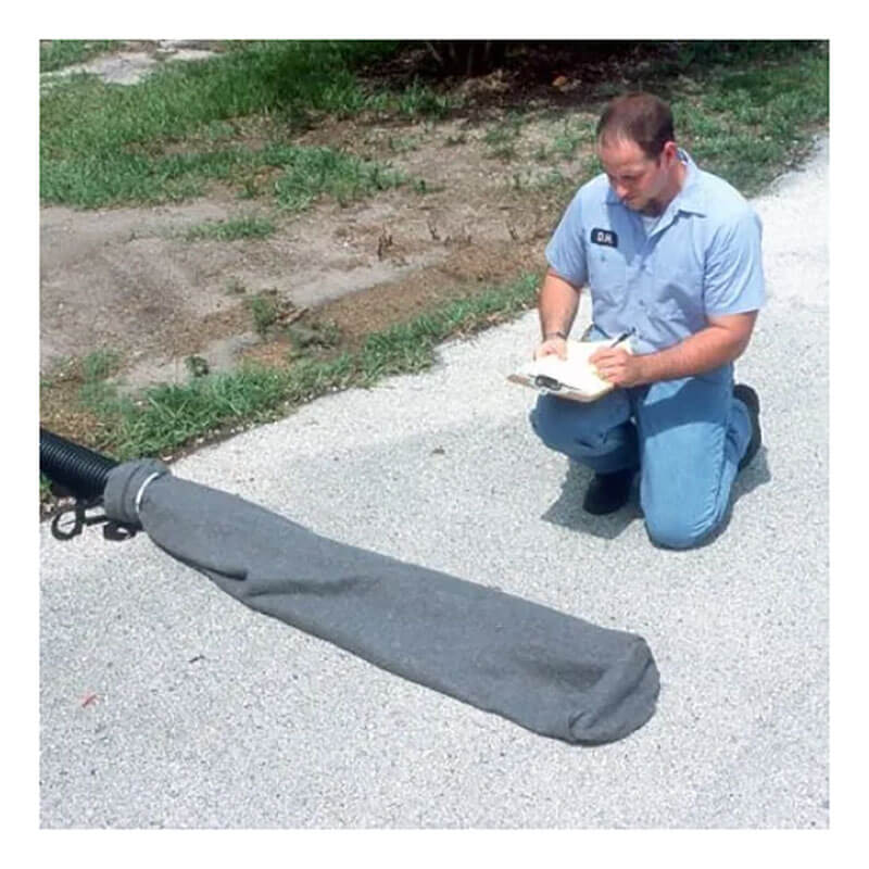 A person in a blue uniform kneels on a paved surface near grass, jotting notes in a notebook. In front is an UltraTech Oil & Sediment Model Pipe Socks 970_-OS, a gray covered object with a black hose for sediment and oil control.