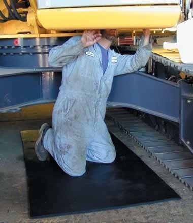 A person in a blue jumpsuit is kneeling on a Working Concepts ErgoKneel Ground Blanket 6060, examining the underside of a large industrial machine with tracks. Their head is partially obscured by the machine.