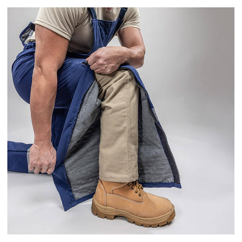 Wearing tan work boots and khaki pants, a person kneels on one knee and unzips the Enespro ArcGuard Performance 40 Cal Arc Flash Bib ARC40B by NSA, revealing the clothing underneath against a neutral background.