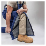 Wearing tan work boots and khaki pants, a person kneels on one knee and unzips the Enespro ArcGuard Performance 40 Cal Arc Flash Bib ARC40B by NSA, revealing the clothing underneath against a neutral background.
