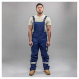 A man stands against a gray background wearing a light T-shirt, tan work boots, and NSA Enespro ArcGuard Performance 40 Cal Arc Flash Bib ARC40B coveralls with reflective leg bands for enhanced arc flash protection.