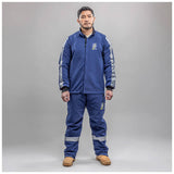 A man faces forward in a blue NSA Enespro ArcGuard Performance 40 Cal Arc Flash Jacket (ARC40J) with reflective sleeve and pant strips, tan work boots, and NSA logos on the chest and leg, set against a plain gray background.