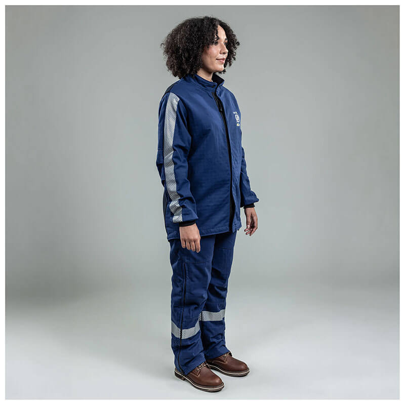 A person with curly hair stands in a studio wearing the NSA Enespro ArcGuard Performance 40 Cal Arc Flash Jacket ARC40J with reflective stripes and brown work boots, facing slightly right against a plain gray background.