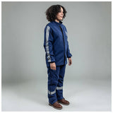 A person with curly hair stands in a studio wearing the NSA Enespro ArcGuard Performance 40 Cal Arc Flash Jacket ARC40J with reflective stripes and brown work boots, facing slightly right against a plain gray background.