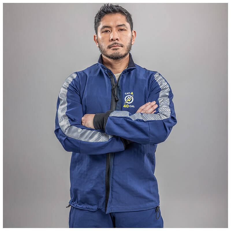 A man with short dark hair and a beard stands with arms crossed, wearing a blue NSA Enespro ArcGuard Performance 40 Cal Arc Flash Jacket (ARC40J) with silver reflective stripes and a "CAT 4 40 CAL" logo, against a plain gray background.