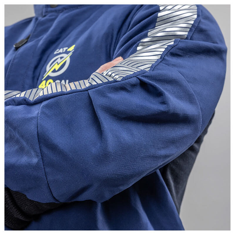 A person wearing an NSA Enespro ArcGuard Performance 40 Cal Arc Flash Jacket (ARC40J) with blue fabric, reflective silver sleeve stripes, and a yellow and white logo on the chest stands with their arms crossed.