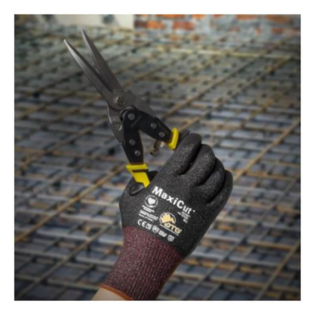 A gloved hand wearing PIP MaxiCut Ultra Nitrile MicroFoam A6 Gloves 44-6745F holds metal snips at a construction site, with steel reinforcing bars visible in the background.