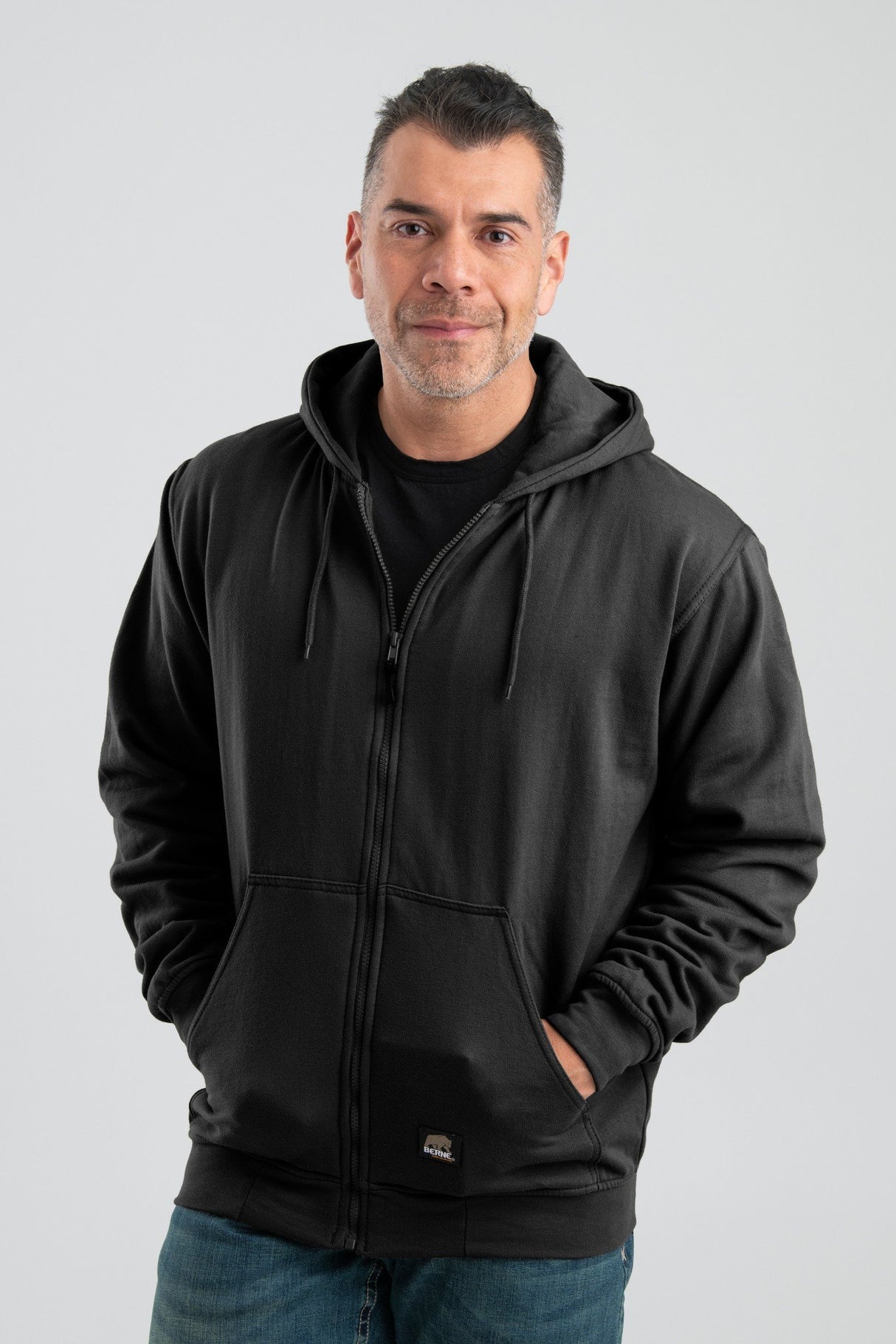 A person wearing a Berne 14 oz Black Thermal Lined Hooded Sweatshirt (model SZ101BK) and blue jeans stands against a neutral background. They have short hair and hands in sweatshirt pockets, with a slight smile on their face.