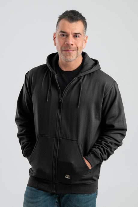 A man is standing with his hands in the pockets of a Berne 14 oz Black Thermal Lined Hooded Sweatshirt SZ101BK. He is smiling and wearing blue jeans against a plain, light-colored background.