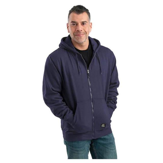 A man stands against a plain white background wearing the Berne 14 oz Navy Thermal Lined Hooded Sweatshirt, his hands comfortably nestled in the oversized front pockets. With short, dark hair and a slight smile, he pairs the look with blue jeans and a black shirt, showcasing a casual winter workwear style.