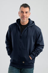 A man wearing the Berne 14 oz Navy Thermal Lined Hooded Sweatshirt (model SZ101NV) and jeans stands against a plain background, smiling with his hands comfortably tucked into the oversized front pockets.