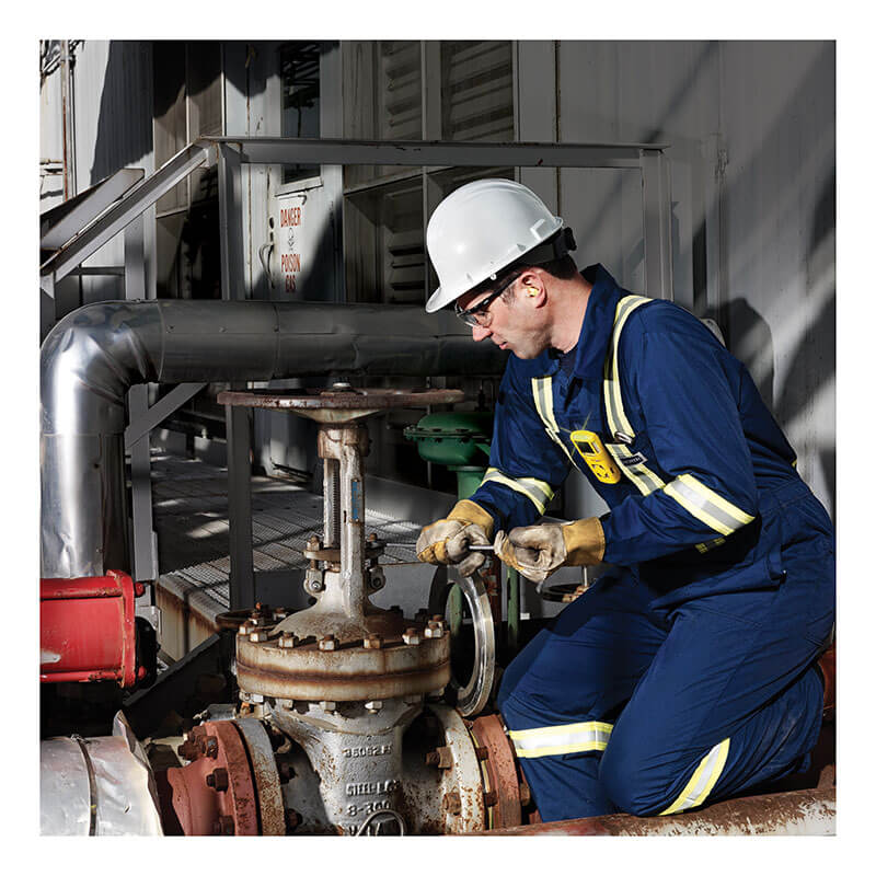 A worker in blue coveralls, safety gear, and a white hard hat uses a BW Technologies by Honeywell GasAlert MicroClip X3 4-Gas Detector MCX3-XWHM-Y-NA to inspect an outdoor industrial valve near metal pipes and machinery.